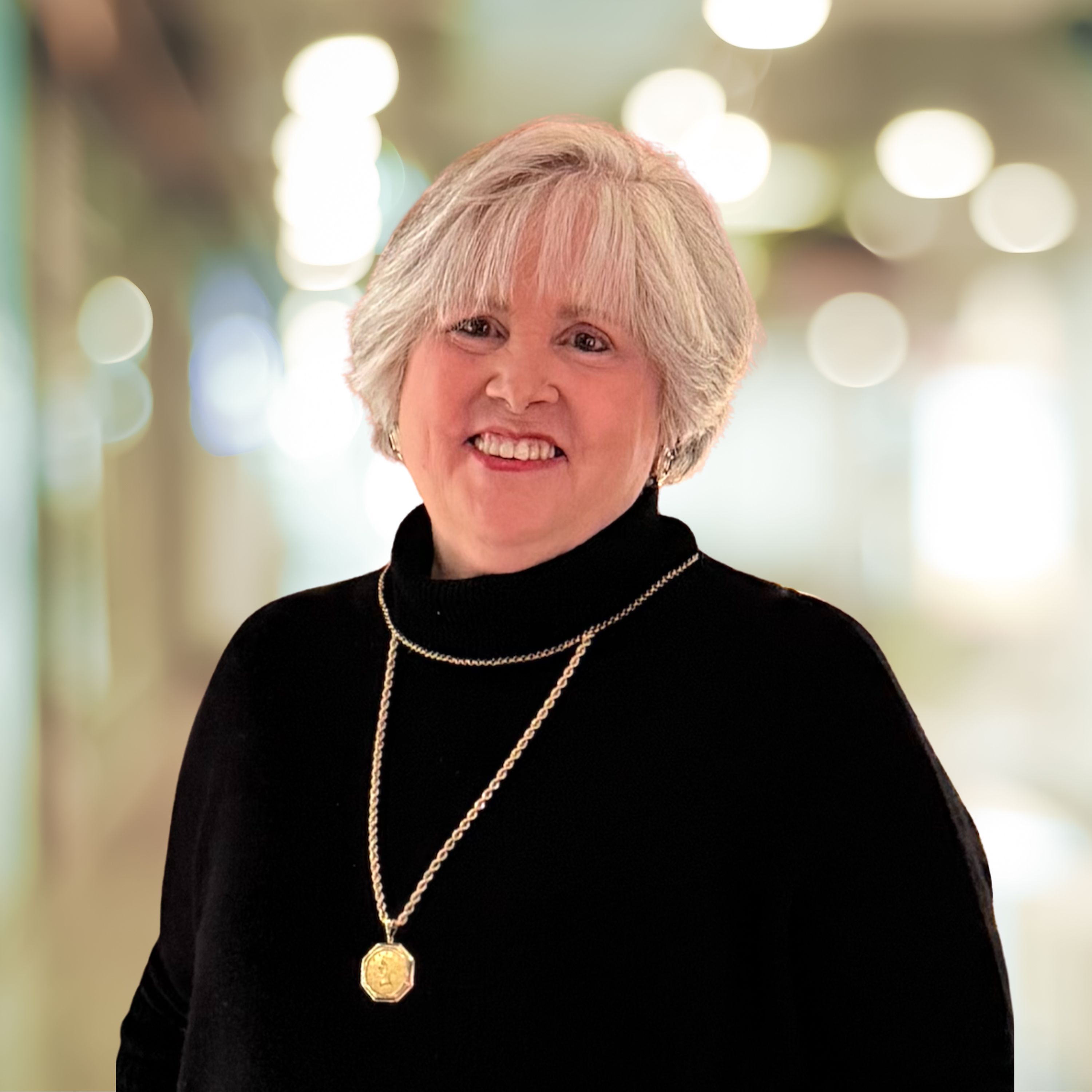 Smiling middle-aged woman with short blonde hair wearing a black top, white textured blazer, and pearl necklace in a well-lit living room.