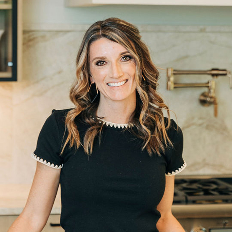 Smiling woman with wavy brown hair and light makeup wearing a black top standing in modern kitchen with beige countertops and appliances