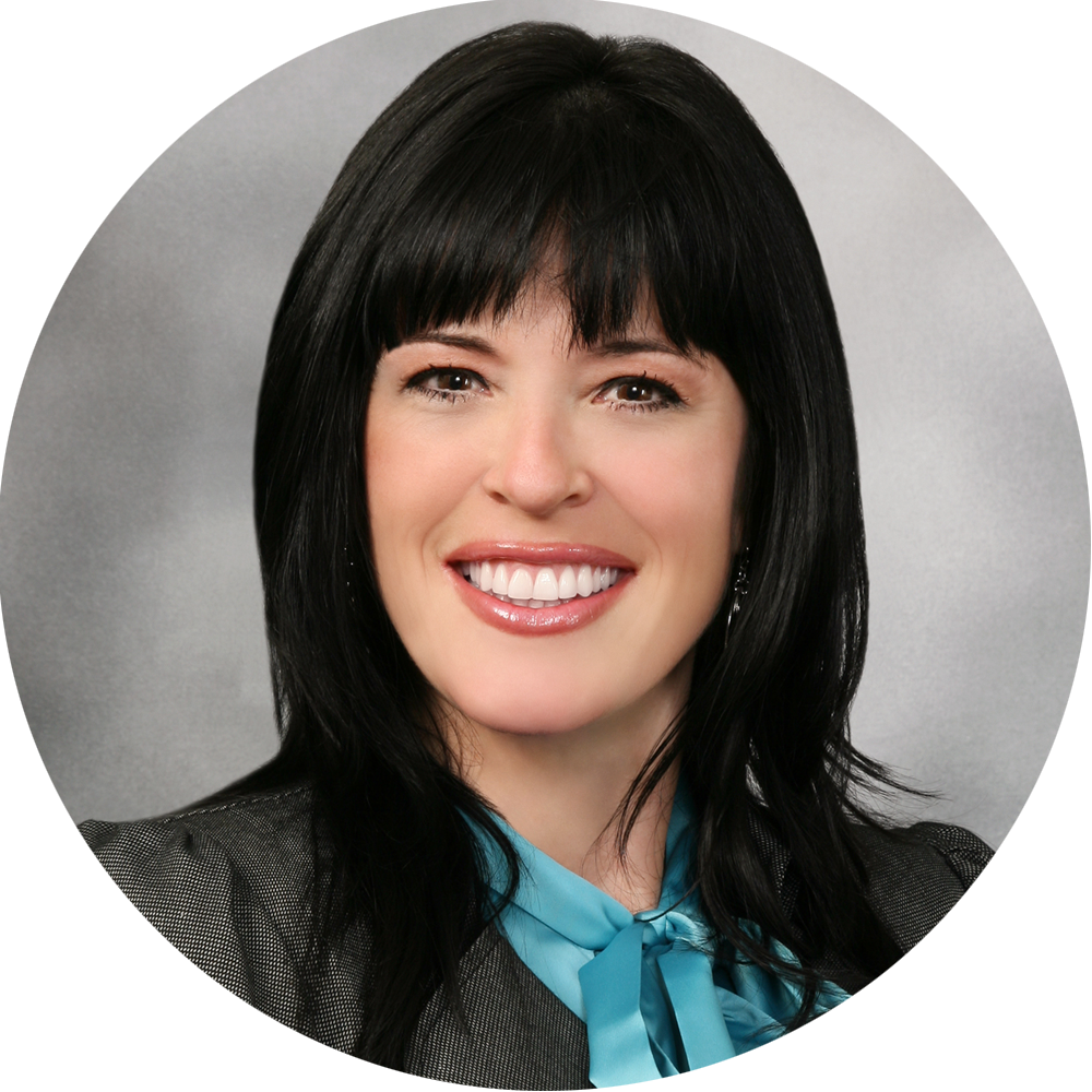 Smiling woman with black hair and bangs wearing a blue blazer against a dark background