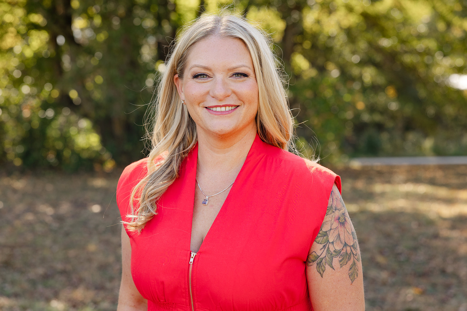 Close-up portrait of a smiling woman with blonde hair and a floral tattoo on her left arm, outdoors in natural light, wearing a sleeveless red-pink top and a silver necklace holding a square-gem.