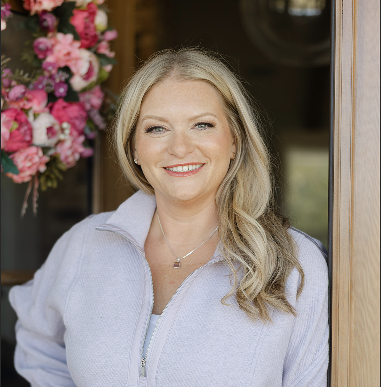 Close-up portrait of a smiling woman with blonde hair and blue eyes, outdoors in natural sunlight, wearing a navy top with white lace collar.
