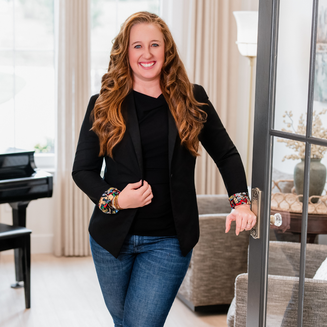 Smiling woman with long curly red hair wearing a black blazer with colorful patterned cuffs, black top, and blue jeans, standing in a bright living room next to a glass door.