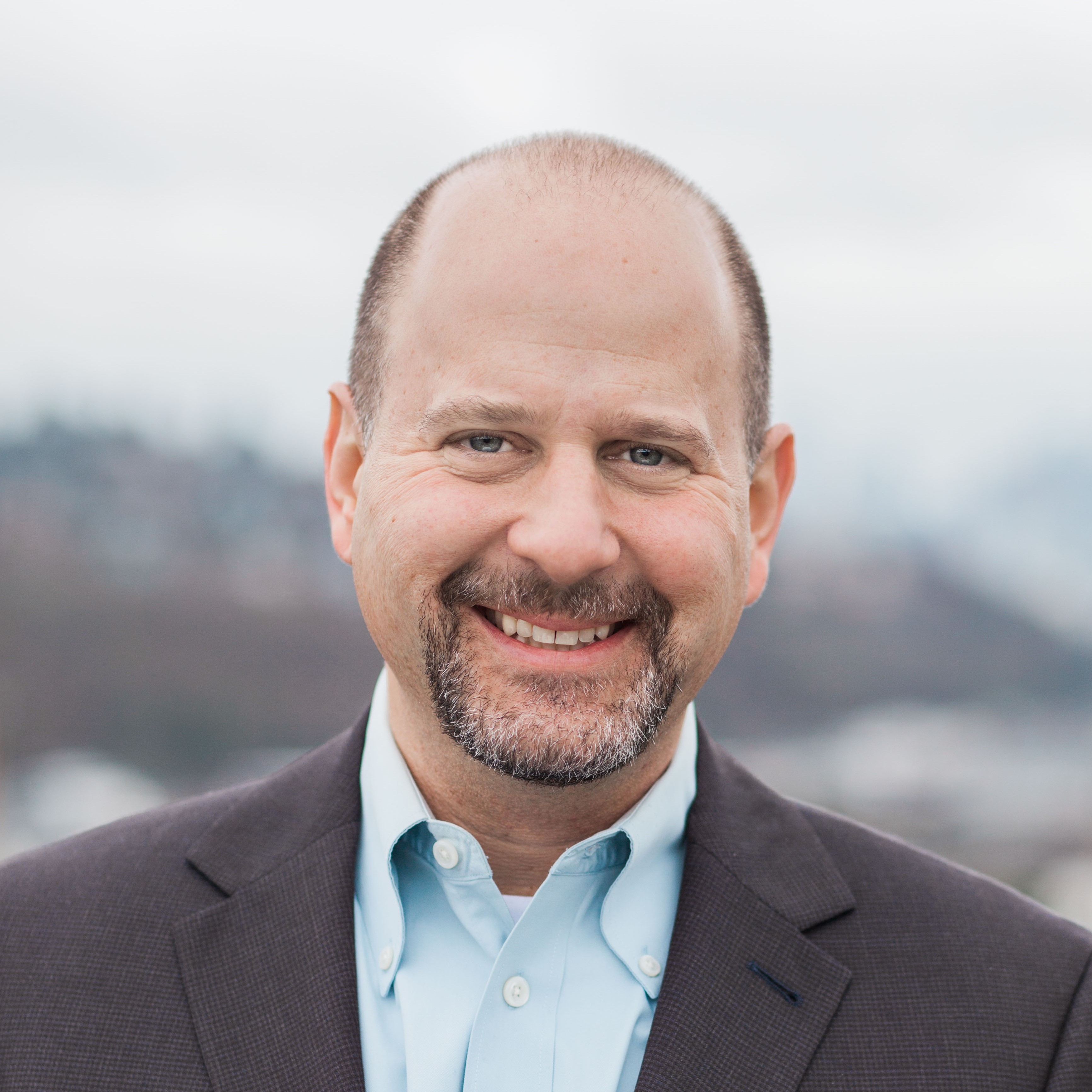Professional headshot of a middle-aged man with a shaved head and goatee, wearing a dark blazer and light blue dress shirt, smiling outdoors with a blurred cityscape background.