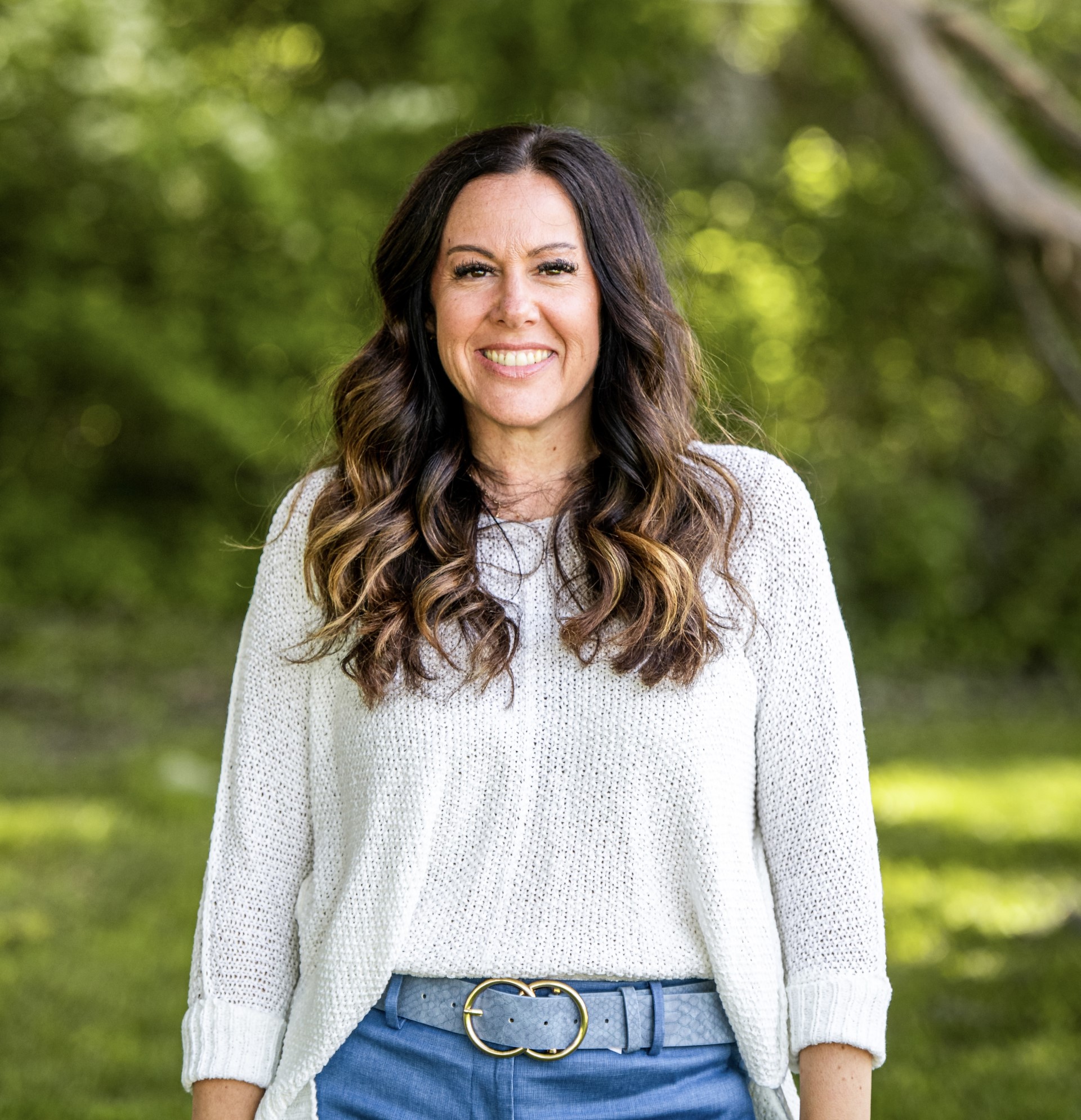 Portrait of a smiling woman with long, wavy brown hair in a white knit sweater and blue pants, standing outdoors in a lush green park.