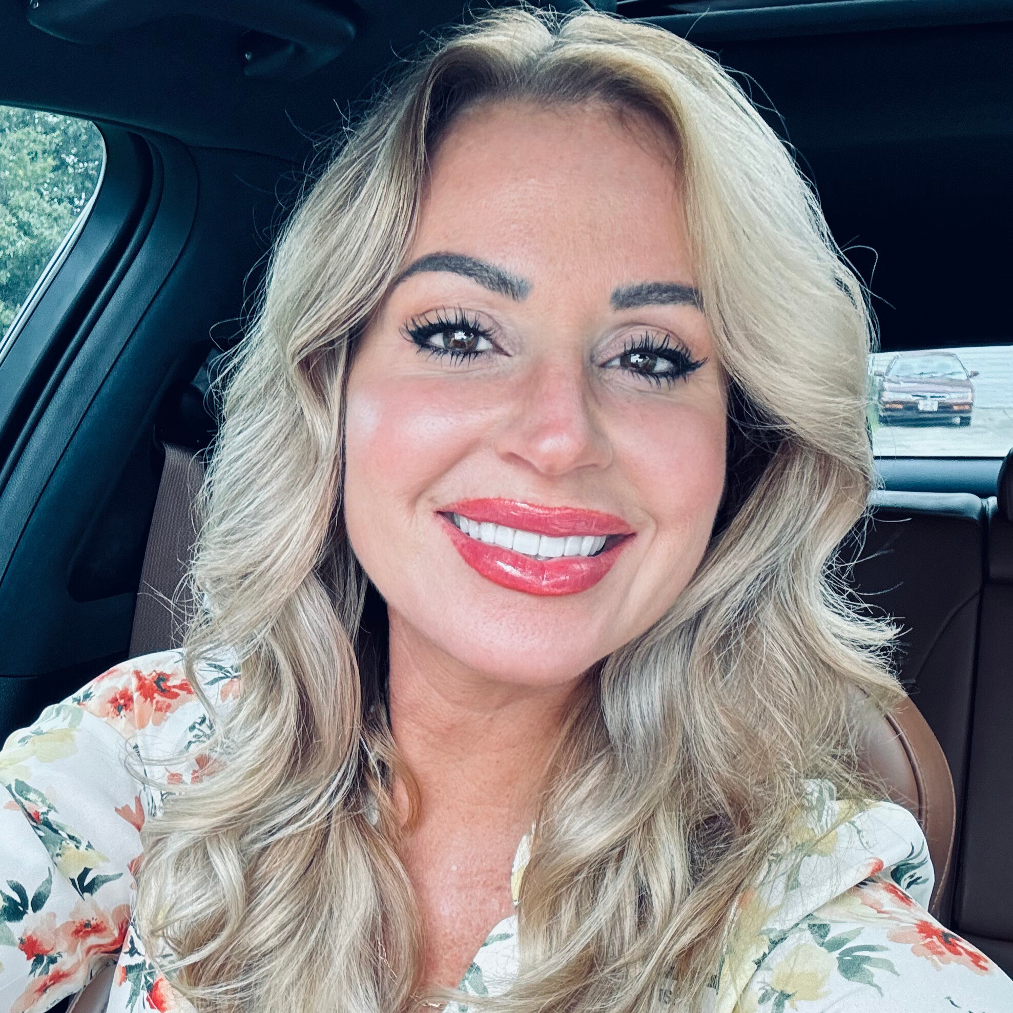 Close-up of a smiling woman with long, wavy blonde hair, wearing bright makeup, sitting inside a car with a parking lot visible in the background.