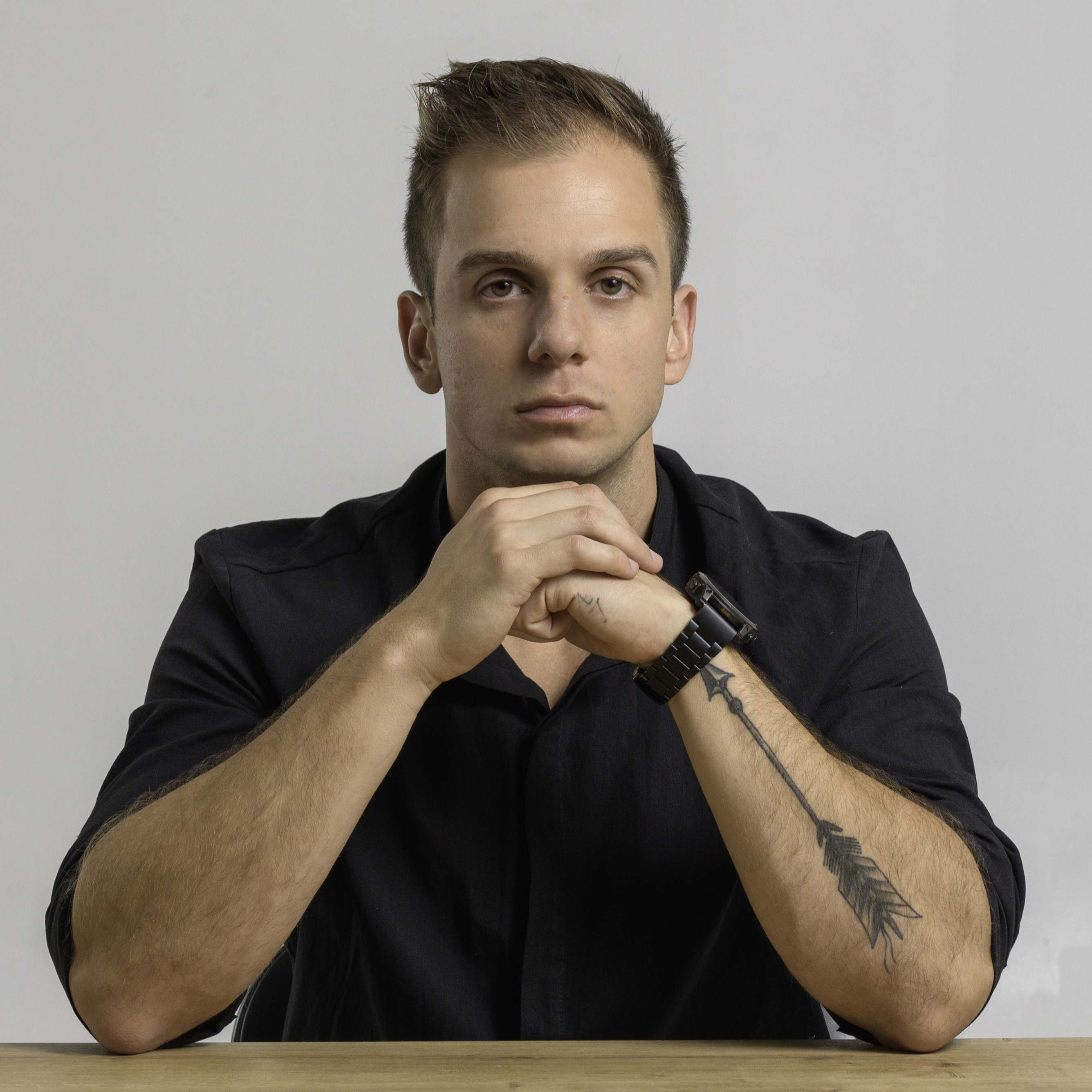 Portrait of a young man with short brown hair and a tattoo of an arrow on his right forearm, sitting at a table with hands clasped in front of his chin, wearing a black shirt and watch against a plain grey background.