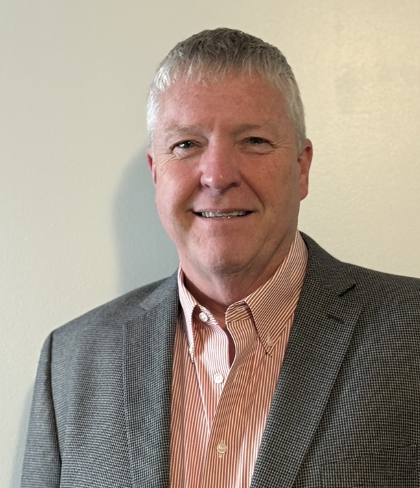 Portrait of a middle-aged man with short gray hair smiling, wearing a gray checkered blazer and a peach and white striped shirt, standing against a plain light-colored background.