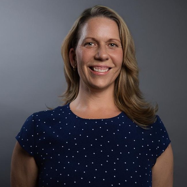 Smiling woman with shoulder-length blonde hair wearing a navy blue polka dot blouse against a gray background