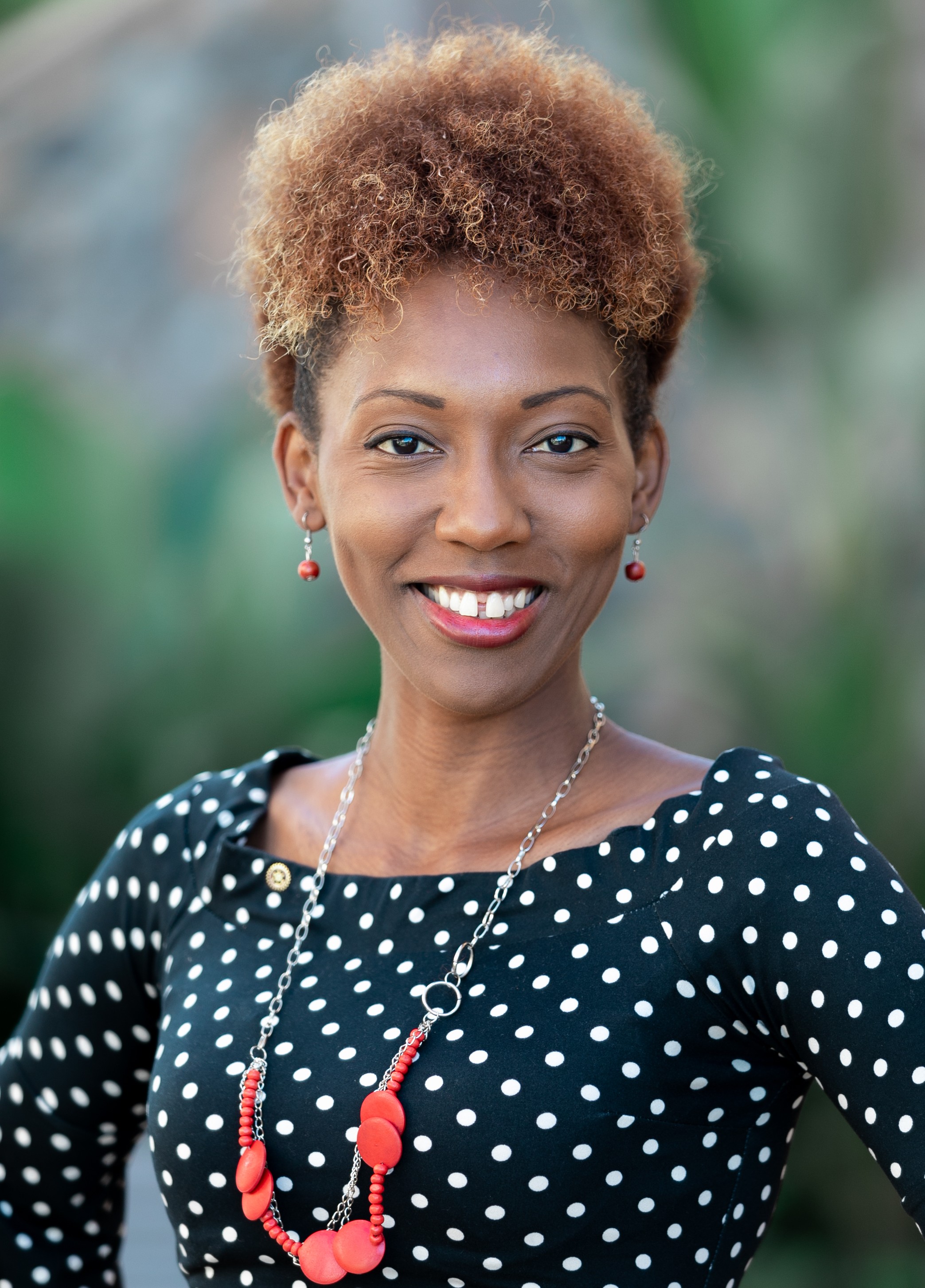 Portrait of a cheerful African American woman wearing a black polka dot dress and red jewelry, outdoors with a blurred natural background.