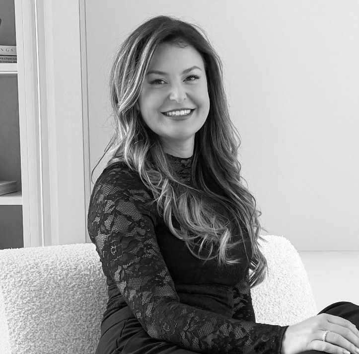 Smiling woman with long wavy hair wearing a black lace top, sitting on a textured armchair in an indoor setting.