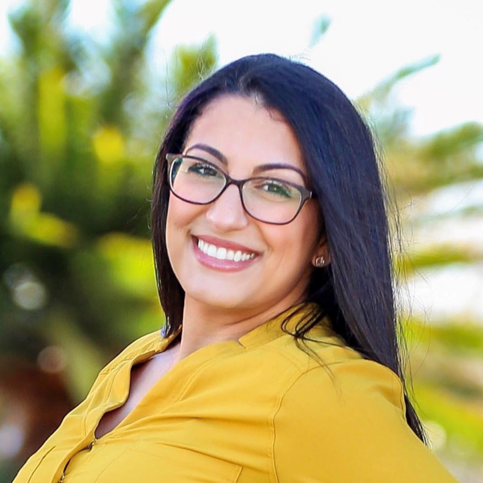 Professional headshot of a smiling man with short black hair and a beard, dressed in a blue blazer and patterned shirt, in a modern interior with decorative elements.