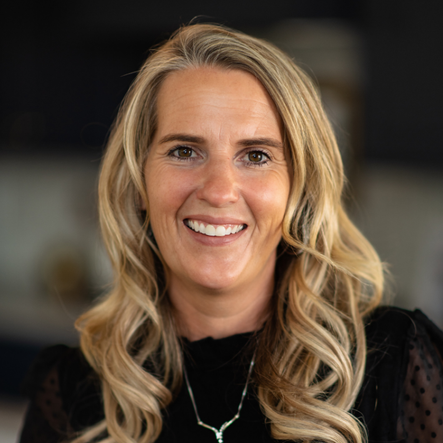 Close-up portrait of a smiling woman with blonde wavy hair, wearing a black top and silver jewelry, in a professional indoor setting
