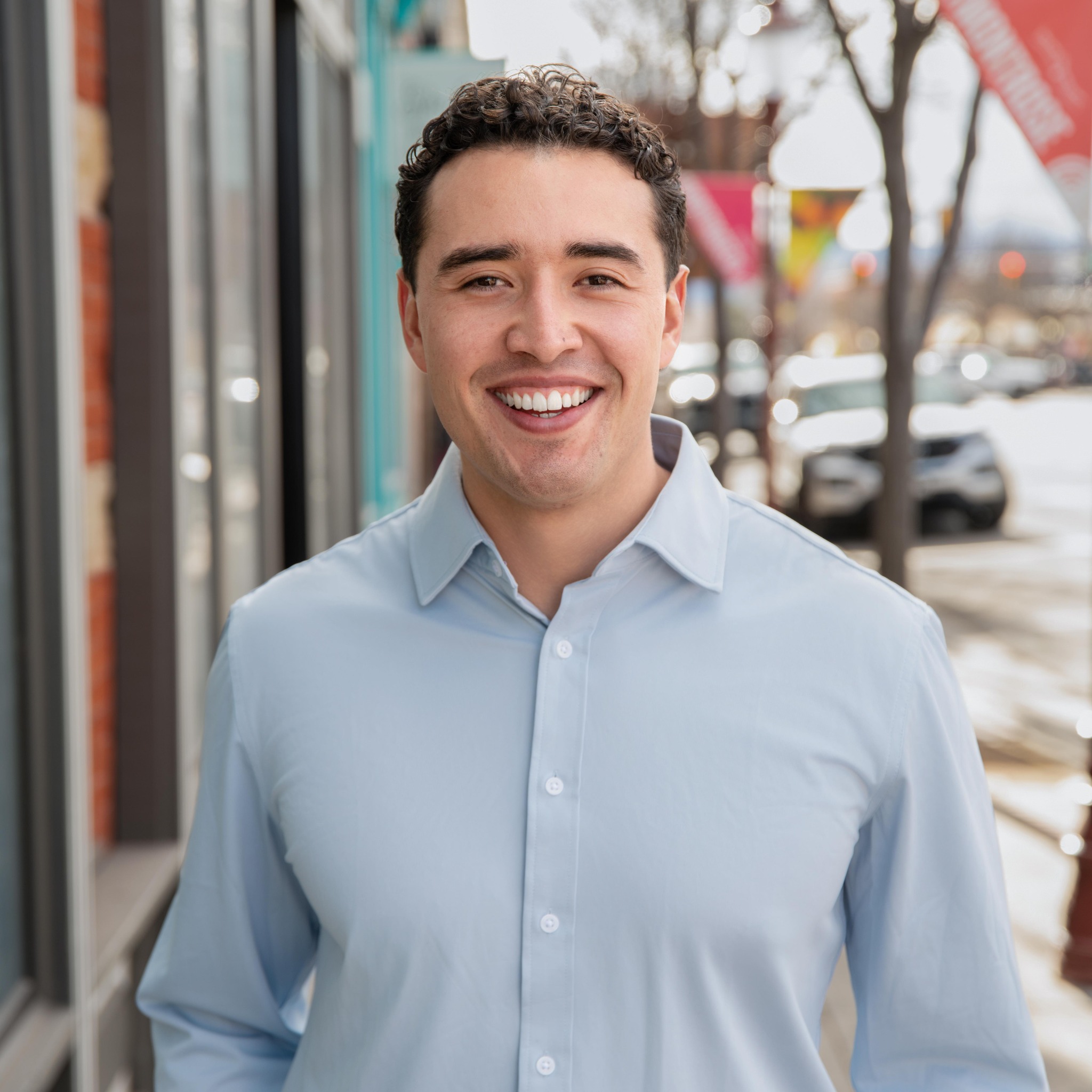 Smiling young man in a light blue dress shirt walking outdoors on a city sidewalk during daytime.